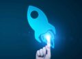A hand pointing at a blue rocket icon on a dark background, symbolizing innovation or technology.