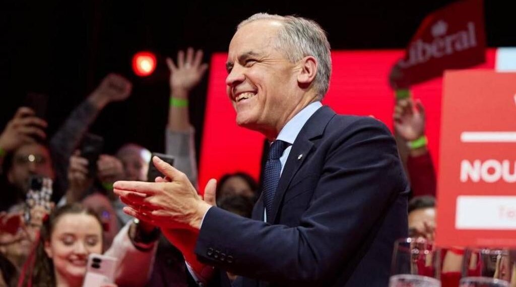 Man in a suit smiling and applauding at a public event with an audience in the background.