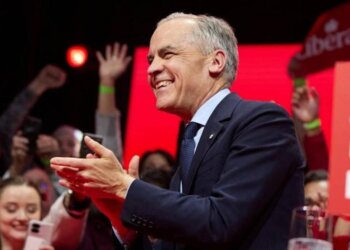 Man in a suit smiling and applauding at a public event with an audience in the background.