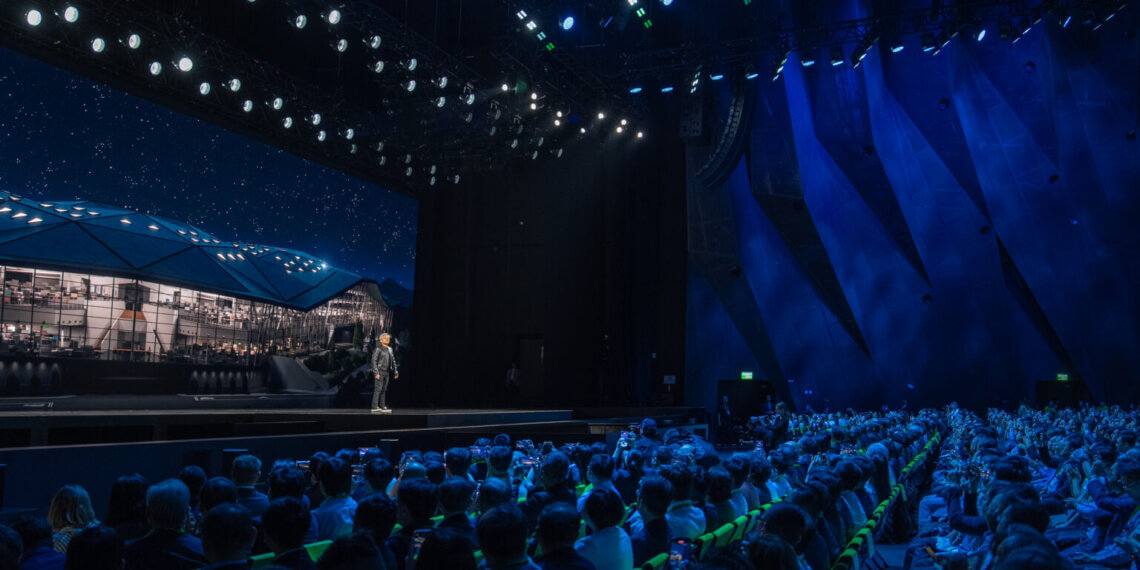 A speaker presenting on stage in front of an audience at a conference event with stage lighting.