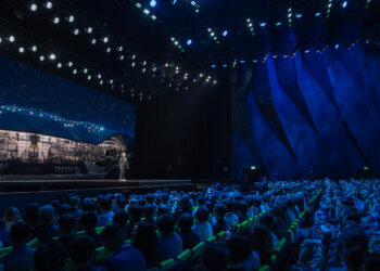 A speaker presenting on stage in front of an audience at a conference event with stage lighting.