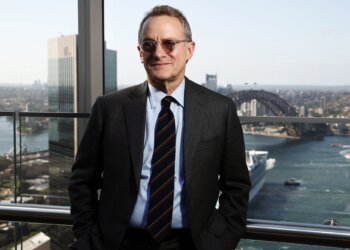 Man wearing glasses and a suit standing in front of Sydney Harbour with the Sydney Harbour Bridge in the background.