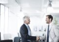 Two men in business attire shaking hands in a modern office environment.