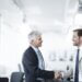 Two men in business attire shaking hands in a modern office environment.