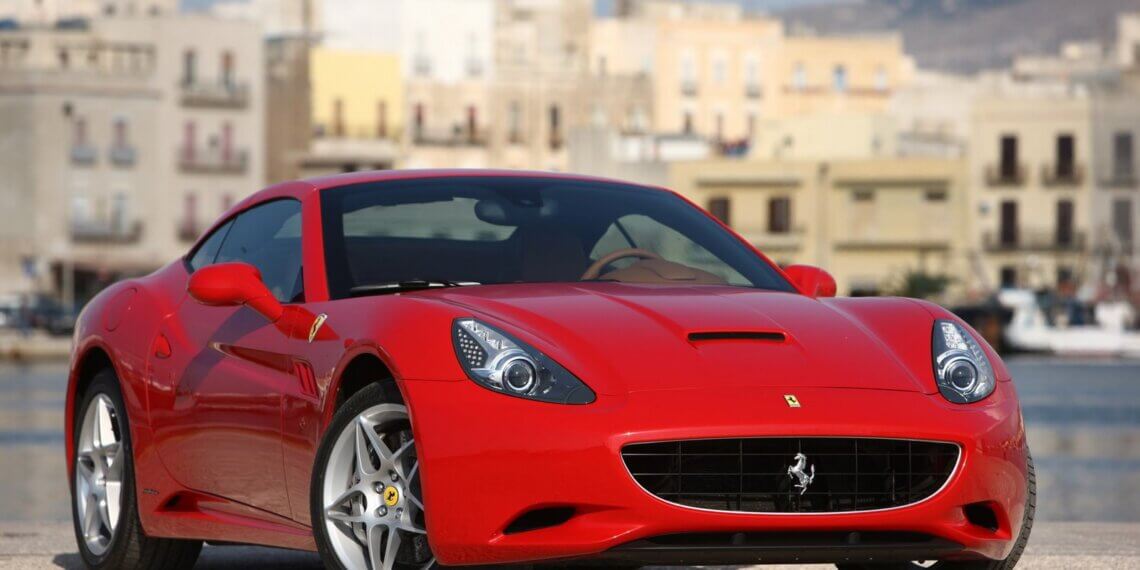 Red Ferrari sports car parked on a paved surface with a coastal background.