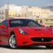 Red Ferrari sports car parked on a paved surface with a coastal background.