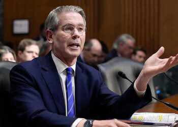 A man in a suit speaking during a Senate hearing, gesturing with his hand while seated at a table.