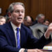 A man in a suit speaking during a Senate hearing, gesturing with his hand while seated at a table.
