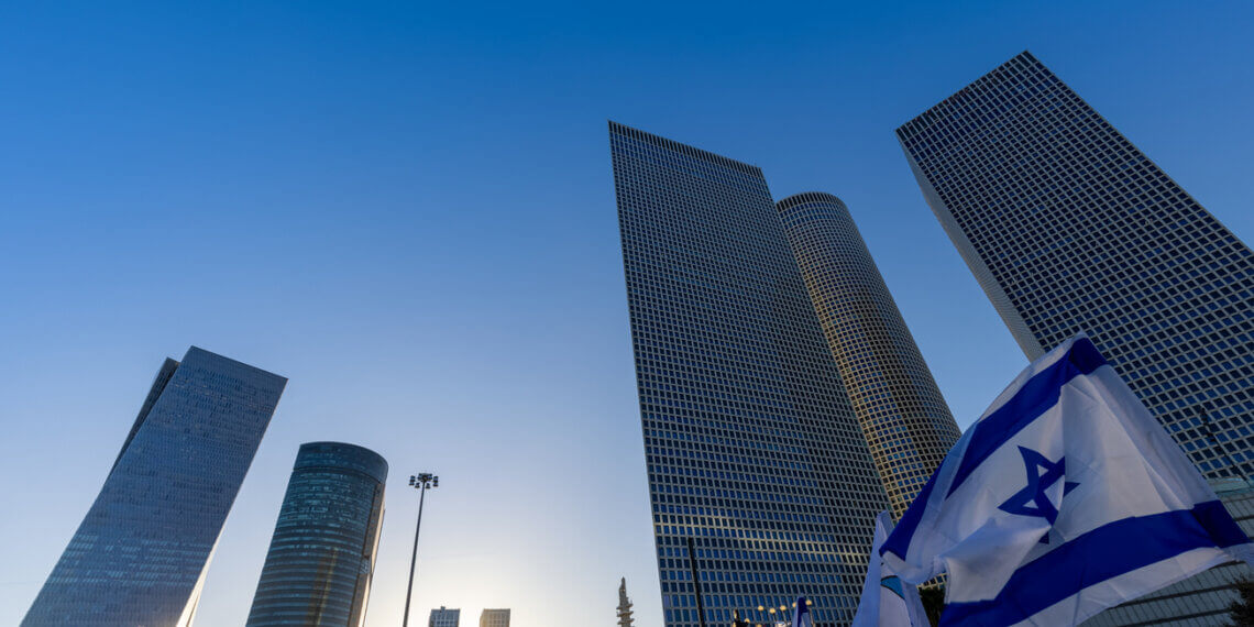 Tall skyscrapers in Tel Aviv with an Israeli flag in the foreground against a clear blue sky.
