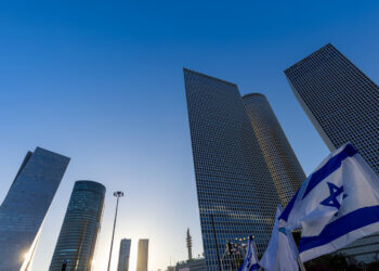 Tall skyscrapers in Tel Aviv with an Israeli flag in the foreground against a clear blue sky.