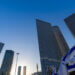 Tall skyscrapers in Tel Aviv with an Israeli flag in the foreground against a clear blue sky.