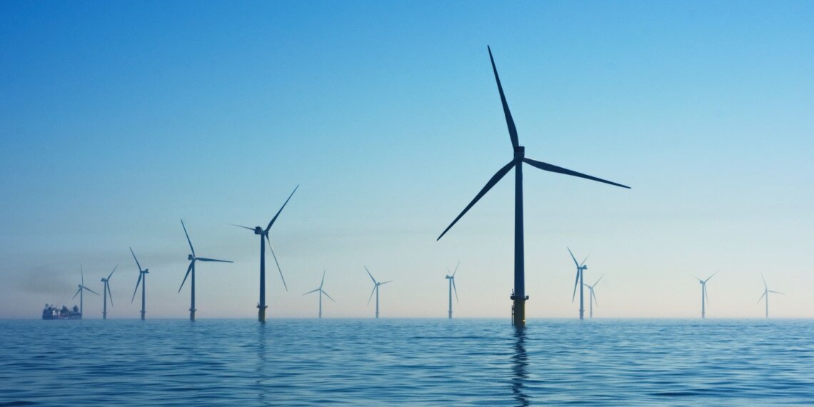 Multiple offshore wind turbines standing in calm water under a clear blue sky.