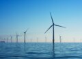 Multiple offshore wind turbines standing in calm water under a clear blue sky.