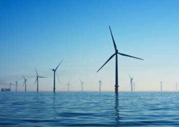 Multiple offshore wind turbines standing in calm water under a clear blue sky.