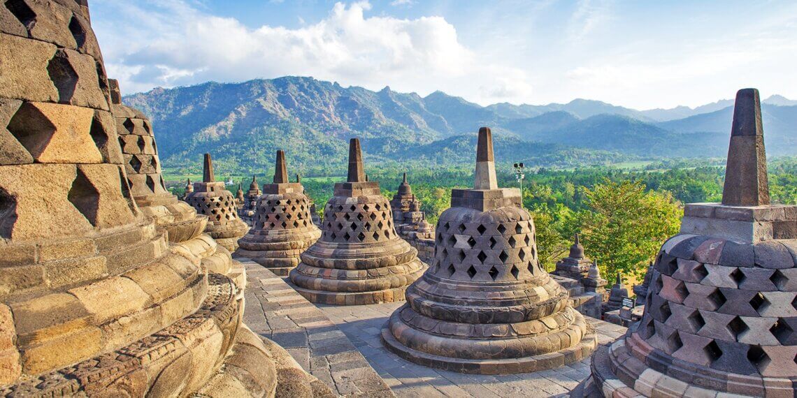 Stone stupas of Borobudur Temple with mountains in the background under a clear sky.