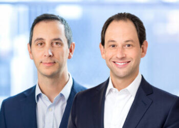 Two men in business suits posing for a portrait, one smiling and the other with a neutral expression.