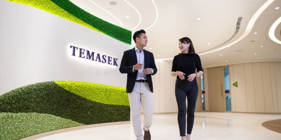Two individuals walking and conversing in a modern office space with a green wall featuring the word 'Temasek'.