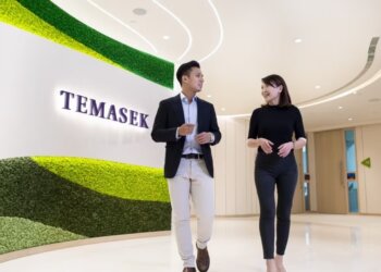 Two individuals walking and conversing in a modern office space with a green wall featuring the word 'Temasek'.
