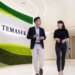 Two individuals walking and conversing in a modern office space with a green wall featuring the word 'Temasek'.