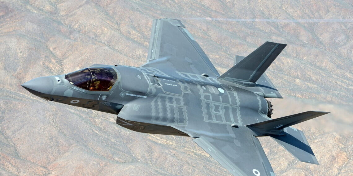 F-35 fighter jet flying in a clear blue sky with a mountainous terrain below.