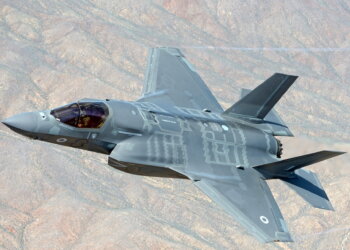 F-35 fighter jet flying in a clear blue sky with a mountainous terrain below.