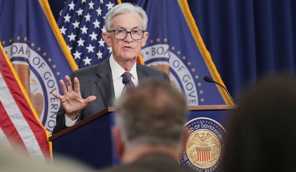 Federal Reserve Chair Jerome Powell addressing an audience during a press conference with flags in the background.