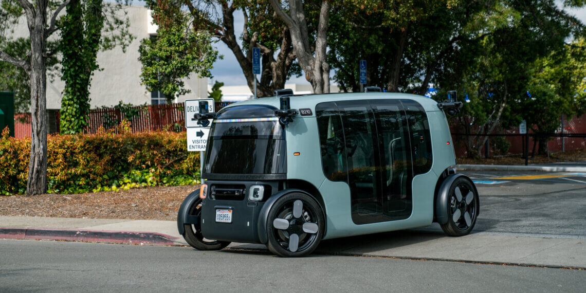 A compact autonomous electric vehicle parked on a street with trees in the background.