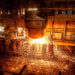 Molten metal being poured from a ladle in an industrial foundry, with sparks flying in the air.