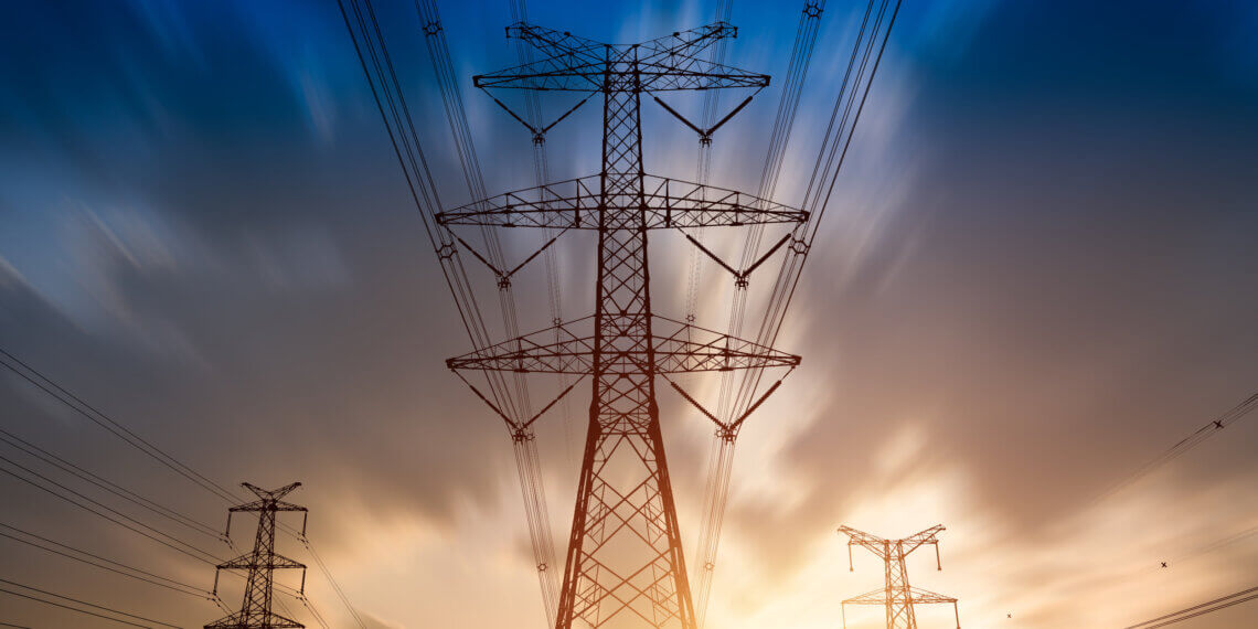 Silhouetted high voltage power lines and towers against a sunset sky with clouds.