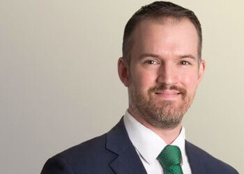 Man wearing a dark suit and a green tie, smiling at the camera with a neutral background.