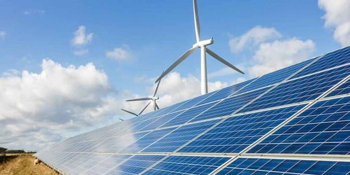 Solar panels arranged in rows with wind turbines visible in the background under a cloudy sky.