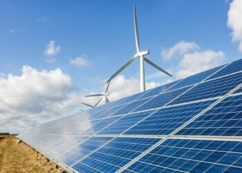 Solar panels arranged in rows with wind turbines visible in the background under a cloudy sky.