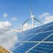 Solar panels arranged in rows with wind turbines visible in the background under a cloudy sky.