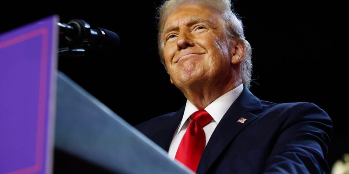 Man in a dark suit and red tie smiling while standing at a podium with a microphone.