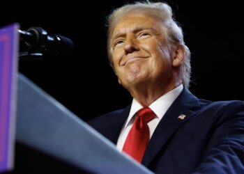 Man in a dark suit and red tie smiling while standing at a podium with a microphone.