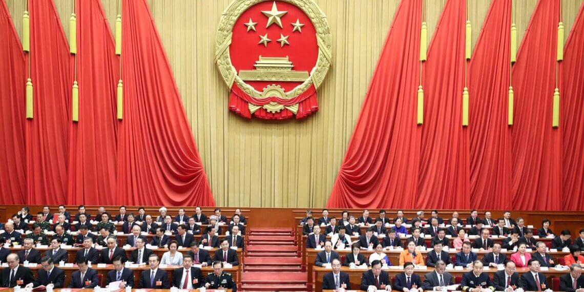Members of the National People's Congress seated in a hall with a large emblem of China in the background.