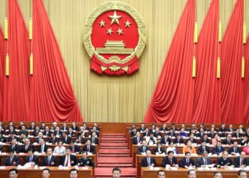 Members of the National People's Congress seated in a hall with a large emblem of China in the background.