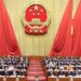 Members of the National People's Congress seated in a hall with a large emblem of China in the background.