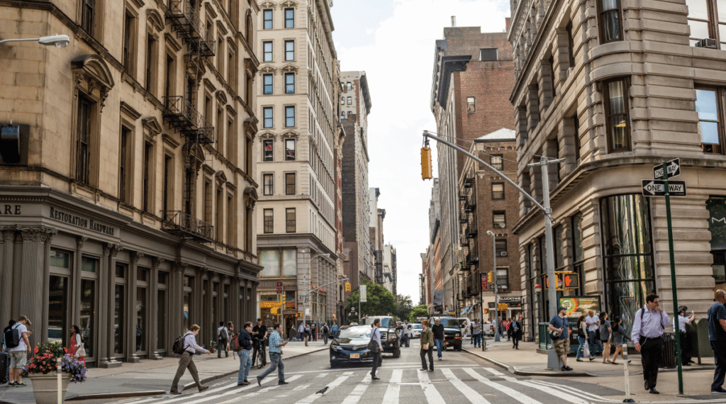 Urban street scene featuring pedestrians crossing at a crosswalk with buildings lining the street.