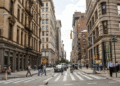 Urban street scene featuring pedestrians crossing at a crosswalk with buildings lining the street.