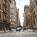 Urban street scene featuring pedestrians crossing at a crosswalk with buildings lining the street.