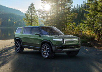 Green Rivian R1S electric SUV parked in a natural setting with trees and sunlight.