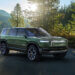 Green Rivian R1S electric SUV parked in a natural setting with trees and sunlight.