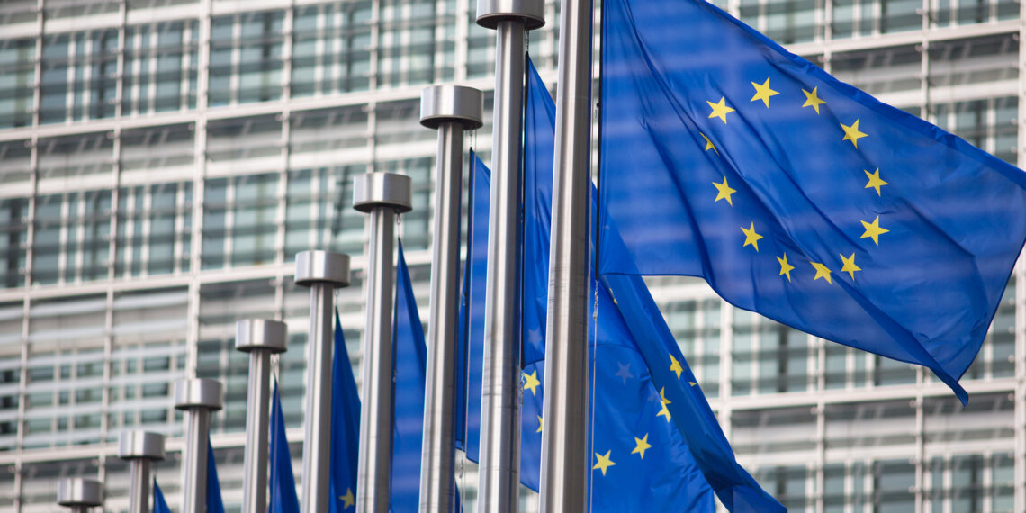 Row of European Union flags waving in front of a modern glass building.