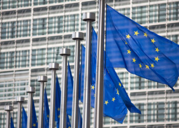 Row of European Union flags waving in front of a modern glass building.