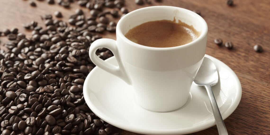 A white coffee cup filled with espresso on a saucer next to scattered coffee beans.
