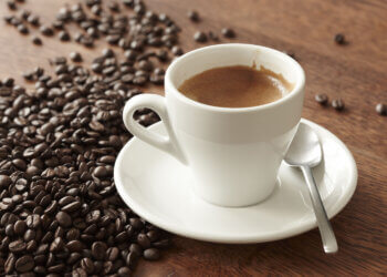 A white coffee cup filled with espresso on a saucer next to scattered coffee beans.