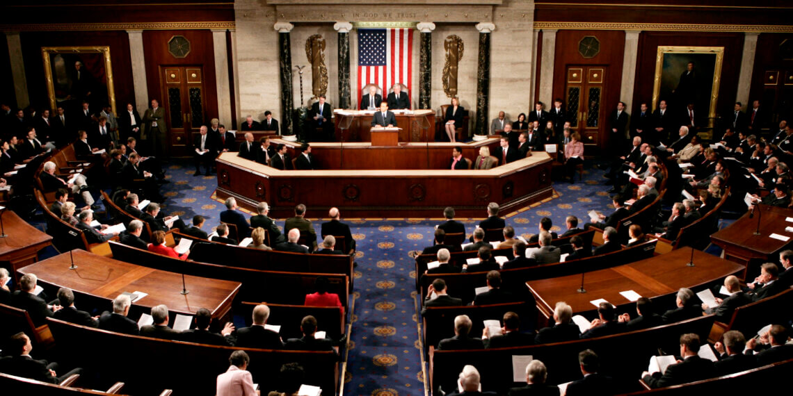 A view of the U.S. Congress chamber with members seated and the American flag displayed.