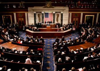 A view of the U.S. Congress chamber with members seated and the American flag displayed.