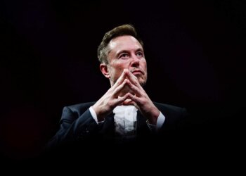 Elon Musk seated with hands clasped, wearing a suit and looking thoughtfully ahead against a dark background.